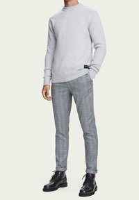 Pull gris à col rond avec poignets côtelés, associé à un pantalon de costume à carreaux dans des tons gris et bleus. Bottes en cuir noir complètent le look.