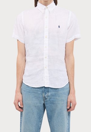 Homme portant une chemise blanche ajustée à manches courtes avec un petit logo brodé bleu marine sur la poitrine et un jean bleu clair.