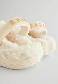 Faux-Fell Hausschuhe in Hellbeige mit einer weichen, flauschigen Textur. Verfügt über einen geformten Riemen mit dem eingravierten Schriftzug 'BAKED' in Hellbraun.