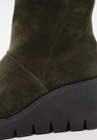 Bottine en daim vert avec une semelle compensée, présentant une finition texturée, des coutures définies et une base plate-forme noire épaisse.