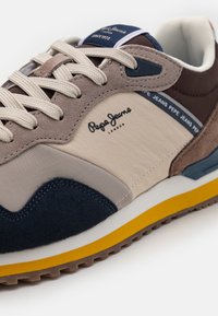 Zapatillas en marrón y beige que combinan materiales de ante y textil, con cordones redondos, acentos en azul, logo distintivo y una suela de goma amarilla.