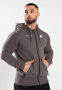 Felpa grigia con zip e dettagli a coste sulle spalle, accenti rossi, toppe con logo e tasche con zip. Realizzata in tessuto testurizzato.