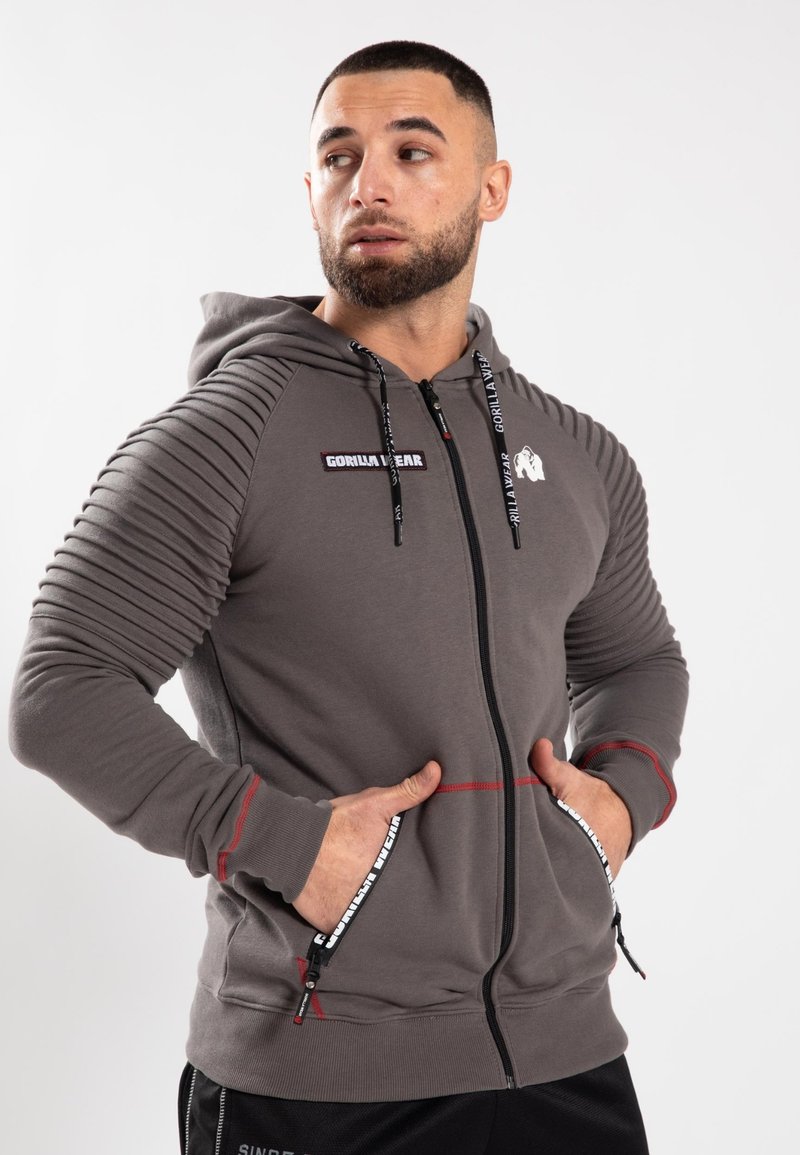 Felpa grigia con zip e dettagli a coste sulle spalle, accenti rossi, toppe con logo e tasche con zip. Realizzata in tessuto testurizzato.