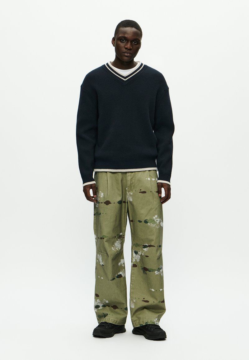 Marineblå strikket sweater med en hvid og grå V-hals. Olivengrønne bukser med malingsplamager. Sorte sneakers. Enkel, afslappet design.