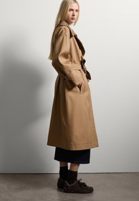 Trench de color marrón claro con cintura ceñida por un cinturón, cuello sobredimensionado y bolsillos frontales. Llevado sobre una falda de mezclilla oscura y zapatillas marrones.