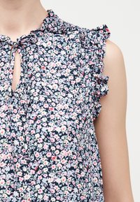 Αμάνικο μπλούζα σκούρου μπλε με μικρά λευκά, ροζ και ανοιχτά γαλάζια λουλούδια, με βολάν στο λαιμό και τις μασχάλες, με δεμένη άνοιγμα στο μπροστινό μέρος.
