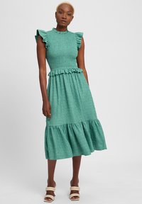 Tee-farbenes Midi-Kleid mit Rüschenärmeln, strukturiertem Stoff, tailliertem Oberteil und gestuftem Rüschensaum. Getragen mit beigen, Riemchensandalen.