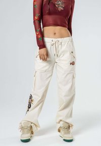 Pantalones cargo beige con diseños florales bordados, cinturilla elástica, bolsillos laterales y corte holgado, combinados con zapatillas de color claro.