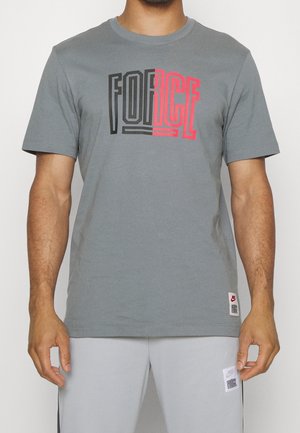 Homme portant un t-shirt gris avec le texte "FORCE" en noir et rouge sur la poitrine, associé à un short gris clair avec un patch logo.