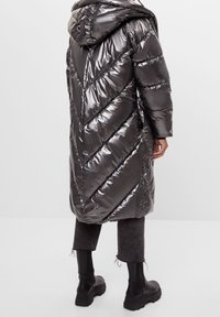 Lång, glänsande mörkgrå puffjacka med huva, med ett diagonalt kviltat mönster, bärs över grå cropped byxor och svarta stövlar.