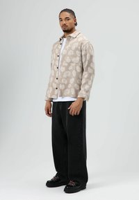 Beige overhemdjack met patroon en zwarte knopen, gedragen over een wit T-shirt. Zwarte, losse broek en stevige zwarte schoenen maken de outfit af.