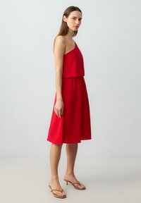 Rode mouwloze jurk met een vloeiende rok, elastische tailleband en een gladde textuur. Gepaarde met beige sandaaltjes met bandjes. Zijaanzicht.