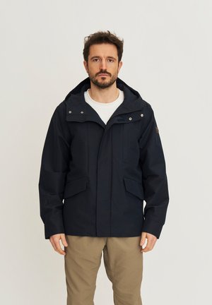 Man draagt een marineblauwe capuchonjas over een wit shirt en beige broek, staand tegen een effen lichte achtergrond.