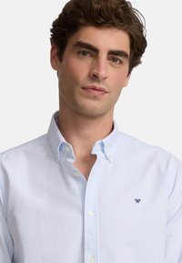 Camisa de rayas azul claro, de botones, hecha de algodón. Cuenta con un cuello, botones frontales y un pequeño logo bordado en el pecho.
