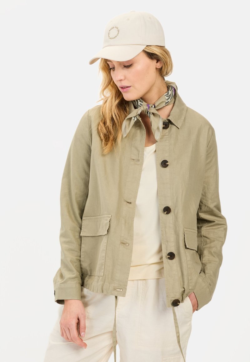 Khaki jakke med knappelukning, to lommer og justerbare snore. Båret over en lys top, parret med en beige baseballcap og et mønstret tørklæde.