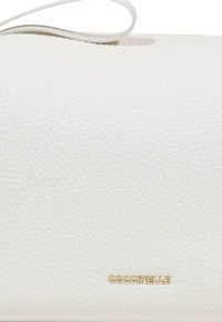 Witte handtas van structuurleer met dunne band en klein gouden "COCCINELLE" logo, gecentreerd vlakbij de onderkant.