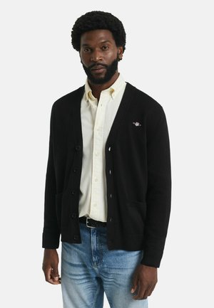 Maglione cardigan nero con scollatura a V, bottoni sul davanti e due tasche frontali, indossato sopra una camicia chiara e jeans blu.