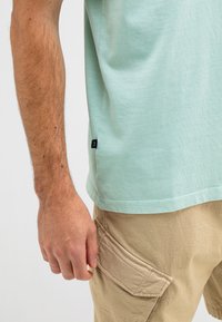 Light green cotton t-shirt with a round neckline, short sleeves, and a black logo tag, paired with khaki cargo shorts featuring side pockets.