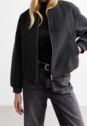 Šedá bomber bunda se zipem vpředu, s žebrovanými manžetami a bočními kapsami, stylizovaná přes černé tričko a spárovaná s tmavými džíny.