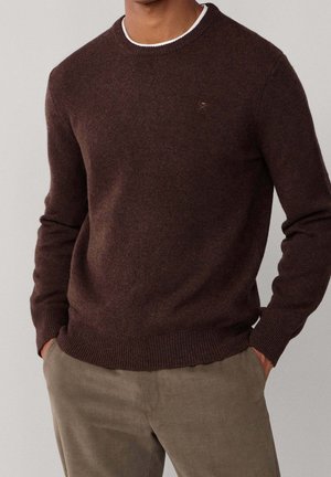 Homme portant un pull marron foncé à col rond par-dessus un sous-vêtement blanc, les mains dans les poches, associé à un pantalon marron clair.