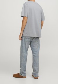 Osoba oblečená v světlých modrých volných džínách, šedé oversized polokošili s krátkým rukávem a hnědých pantoflích, stojící na jednobarevném pozadí.