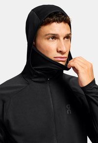 Schwarzer Performance-Hoodie mit hohem Kragen und angenählter Kapuze. Texturiertes Material mit einem frontalen Reißverschluss und dezentem Logo auf der linken Brust.