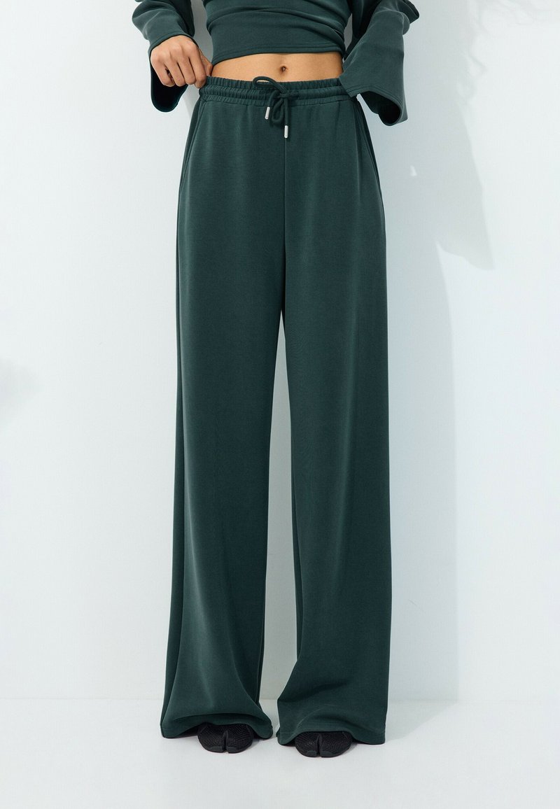 Personne portant un pantalon de survêtement large vert foncé avec taille à cordon et chaussures noires, debout devant un fond blanc uni.