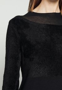 Gros plan d'une personne portant un pull noir texturé et duveteux avec un col noir transparent et des manches longues.
