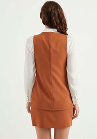 Brun ärmlös klänning över en vit långärmad skjorta; slät vadderad tyg med rak skärning; har knappar på skjortans manschetter.