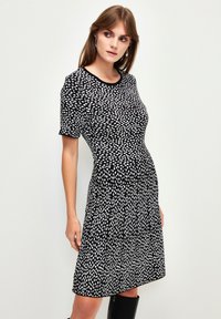 Schwarzes Kleid mit kurzen Ärmeln, das ein weißes gepunktetes Muster aufweist. Der Stoff hat eine glatte Textur und eine taillierte Form im Oberkörper.