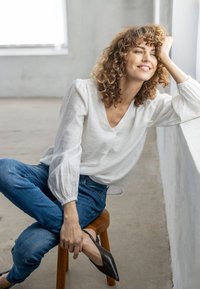 Femme aux cheveux bouclés portant un chemisier blanc et un jean, assise sur un tabouret en bois, souriante et appuyée sur un rebord de fenêtre blanc dans une pièce lumineuse.