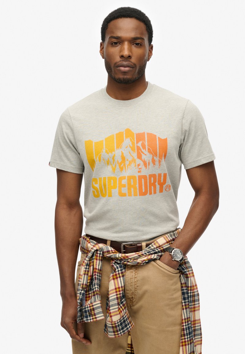 Homme portant un t-shirt Superdry beige avec un motif de montagne orange, une chemise à carreaux nouée autour de la taille, un pantalon beige et une montre au poignet, debout devant un fond blanc.
