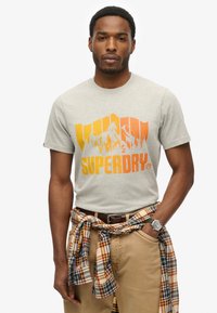 Lys grå bomulds T-shirt med en gradient grafik af orange og gul bjerg samt "SUPERDRY" logo. Parret med brune bukser og en ternet skjorte knyttet om taljen.