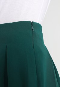 Zelená plisovaná sukně z texturované látky, s decentním bočním zipovým uzávěrem a hladkým pasem.