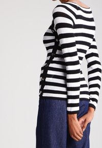 Suéter de punto a rayas en blanco y negro con mangas largas, que presenta detalles de botones a lo largo del costado y una silueta ajustada.