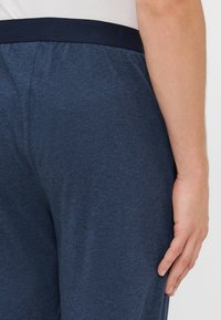 Rechterzij aanzicht van een persoon in donkerblauwe broek met een zwarte tailleband en een wit shirt, hand ontspannen langs hun zij.