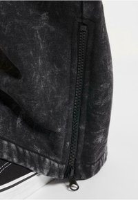 Svart denim tyg med en marmorerad struktur, featuring en svart dragkedjedetalj på sidan, och en avslappnad manschett i botten.