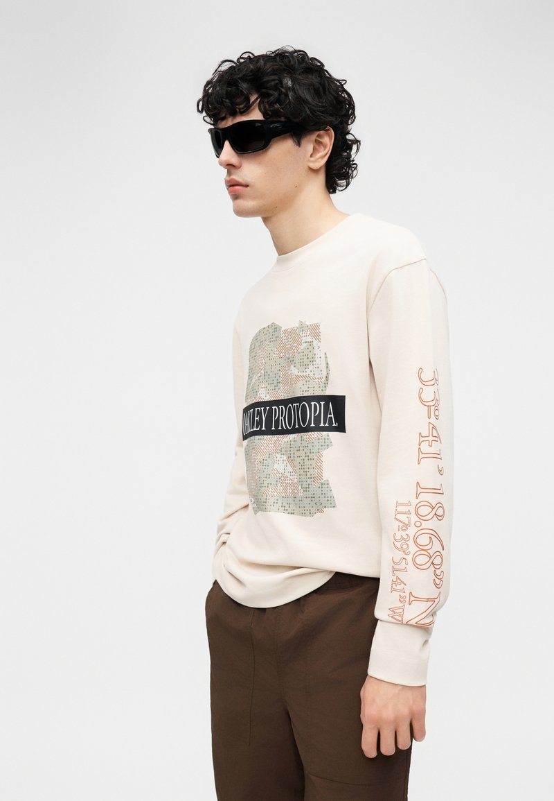 Man with curly hair wearing black sunglasses, beige sweatshirt with graphic and text, brown pants, standing with one hand in pocket.