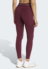 Leggings granate con una cintura alta, textura suave y una franja clara contrastante en la parte superior, combinados con zapatillas deportivas blancas.