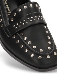 Mocasín de cuero negro con punta redondeada, adornado con tachuelas plateadas y una correa decorativa en la parte superior. Textura suave y acentos metálicos.