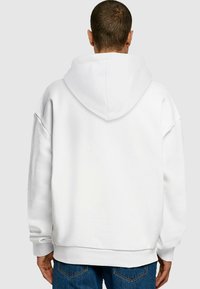 Felpa bianca con cappuccio regolabile, polsini a costine e vestibilità ampia. Il tessuto presenta una texture liscia e un colore uniforme.