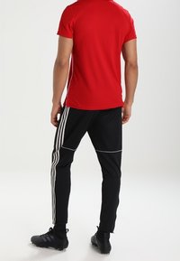 Rotes T-Shirt mit kurzen Ärmeln, kombiniert mit schwarzen, schmal zulaufenden Hosen mit weißen Seitenstreifen. Das Outfit wird durch schwarze Fußballschuhe ergänzt.