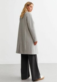 Ljusgrå, knälång kappa med rak skärning och sidslitsar, kombinerad med mörka vida byxor och skor med vit sula.