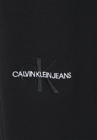 Czarna bawełniana tkanina z haftowanym logo w białym i szarym kolorze. Logo brzmi "CALVIN KLEIN JEANS" z stylizowanym "CK."