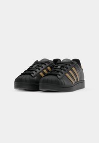 Black sneakers with perforated leather, rubber toe cap, and three brown stripes. Flat sole and lace-up design. Subtle branding on tongue.