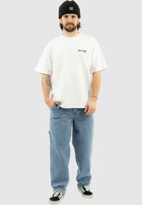 Hvid T-shirt med kortærmer og et lille sort logo, parret med lyseblå løse jeans og sorte Vans sneakers. Sort beanie båret på hovedet.