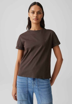 Frau mit langen dunklen Haaren trägt ein einfaches dunkelbraunes T-Shirt und hellblaue Jeans, steht vor einem neutralen grauen Hintergrund.