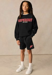 Černá bavlněná mikina se červeným logem "SUPERDRY", kombinovaná s černými šortkami s červeným logem a bílými sportovními ponožkami s bílými teniskami.