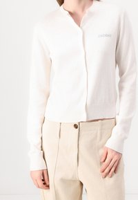 Světle béžový kardigan s kulatým výstřihem, žebrovanými manžetami, knoflíky vpředu a malým vyšitým logem "2ND DAY" na levé části hrudi.