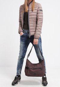 Person i beige puffjacka, svart skjorta, blå jeans, metalliska sneakers, som håller en brun axelväska i remmen mot en vit bakgrund.